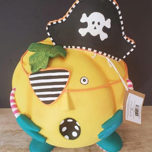 May include: A quirky yellow pumpkin ornament wearing a pirate hat with a skull and crossbones. The pumpkin has a striped eye patch, teal feet, and a leaf. The design includes a dotted pattern and a tag.