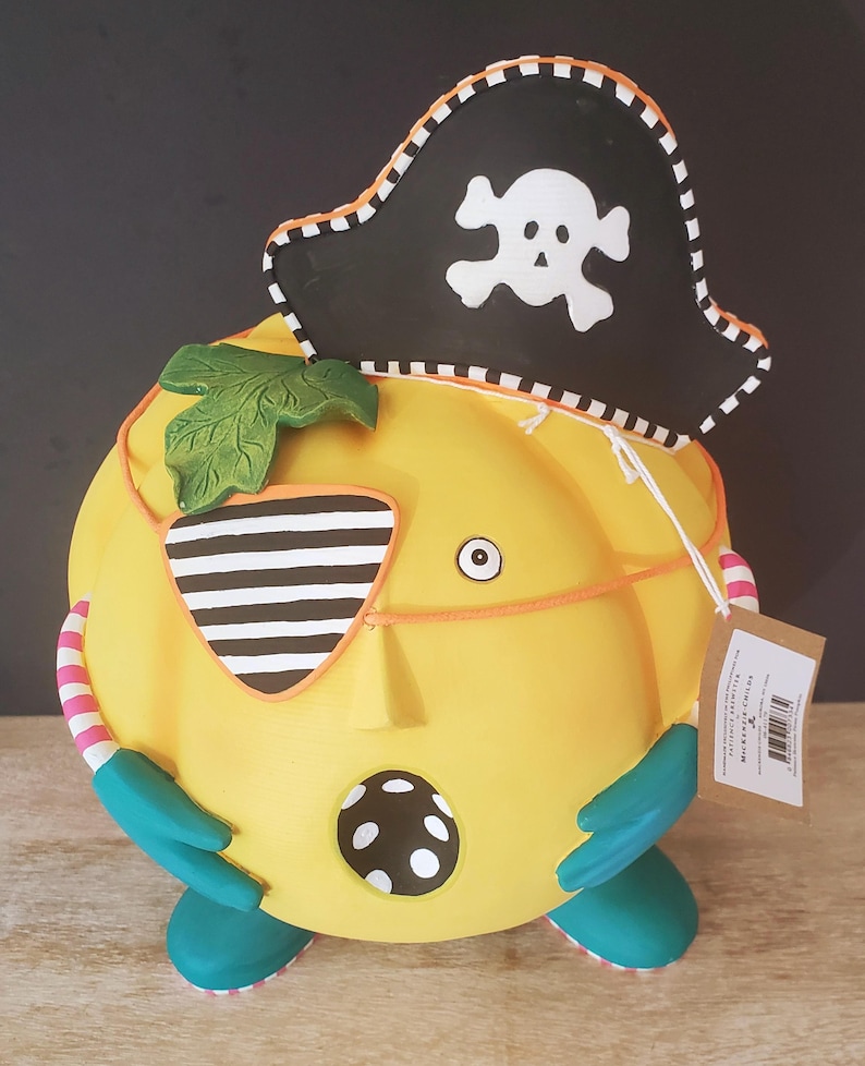 May include: A quirky yellow pumpkin ornament wearing a pirate hat with a skull and crossbones. The pumpkin has a striped eye patch, teal feet, and a leaf. The design includes a dotted pattern and a tag.