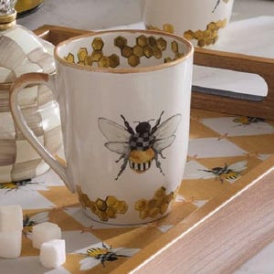 May include: A white ceramic mug featuring a central illustration of a bee with a black and white checkered body and yellow abdomen. The mug is decorated with gold-toned honeycomb patterns around the rim and base, with a matching handle.