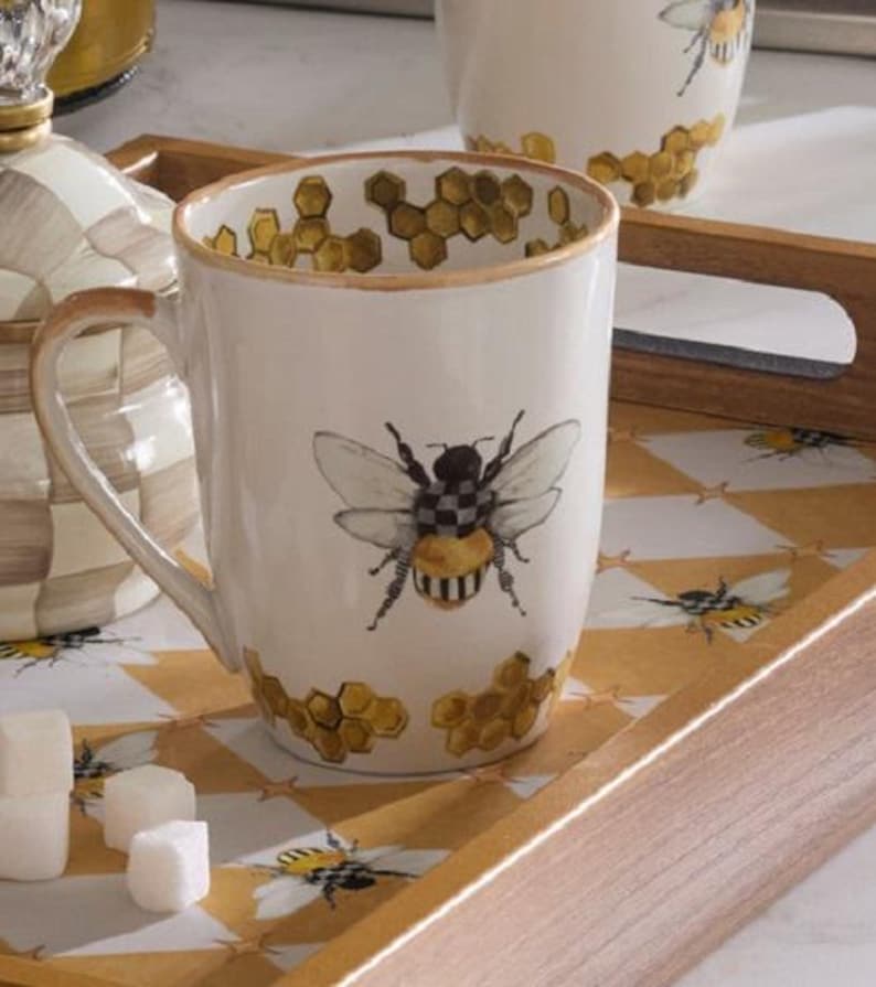 May include: A white ceramic mug featuring a central illustration of a bee with a black and white checkered body and yellow abdomen. The mug is decorated with gold-toned honeycomb patterns around the rim and base, with a matching handle.