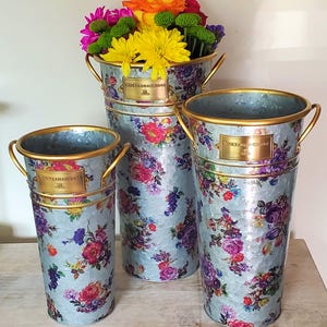 May include: Three decorative metal flower buckets with floral patterns. The buckets are in varying sizes, with gold handles and trim. The largest bucket holds a colorful arrangement of flowers. Each bucket has a gold label that reads "MACKENZIE-CHILDS".