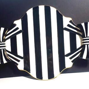 May include: A ceramic serving tray featuring a bold black and white vertical striped pattern. The tray has a scalloped edge with a thin gold rim and is accented on both sides by large, three-dimensional decorative bows in matching stripes.