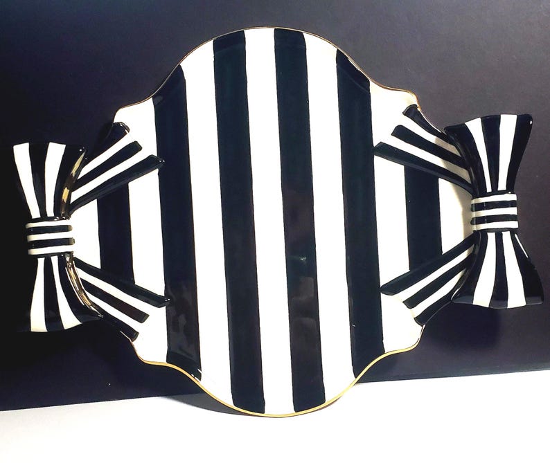 May include: A ceramic serving tray featuring a bold black and white vertical striped pattern. The tray has a scalloped edge with a thin gold rim and is accented on both sides by large, three-dimensional decorative bows in matching stripes.