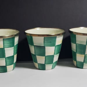 K&ouml;nnte beinhalten: Drei konische Becher mit einem handgemalten gr&uuml;n-wei&szlig;en Karomuster. Jeder Becher hat eine glatte, cremefarbene Innenseite und einen metallisch goldfarbenen Rand an der Oberseite. Die Becher sind in einer horizontalen Reihe vor einem dunklen Hintergrund.