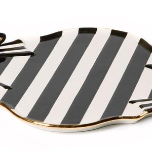 May include: A ceramic serving tray featuring a black and white striped pattern across the surface. The tray has a scalloped edge with a gold-toned trim and is accented with a three-dimensional black and white striped bow on each side.