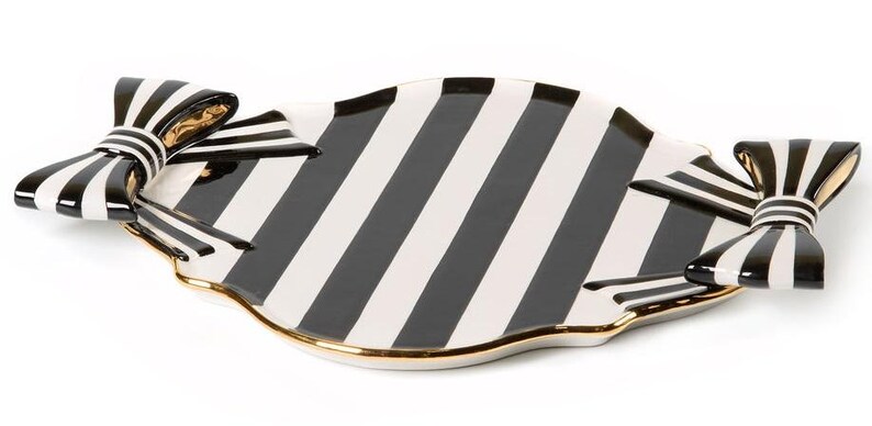 May include: A ceramic serving tray featuring a black and white striped pattern across the surface. The tray has a scalloped edge with a gold-toned trim and is accented with a three-dimensional black and white striped bow on each side.