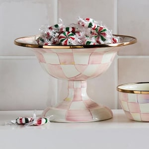 May include: A pink and white checkered pedestal bowl with a gold rim, filled with wrapped peppermint candies. The bowl has a wide, shallow top and a flared base. Two wrapped candies are in the foreground. The bowl is a decorative item.