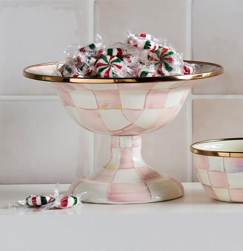 May include: A pink and white checkered pedestal bowl with a gold rim, filled with wrapped peppermint candies. The bowl has a wide, shallow top and a flared base. Two wrapped candies are in the foreground. The bowl is a decorative item.