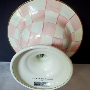 May include: A white and pink checkered cake stand with a gold rim. The base is white with the text "MacKenzie-Childs" and "Made in China / Fabriqu&eacute; en Chine". The stand is designed to hold a cake or other desserts.