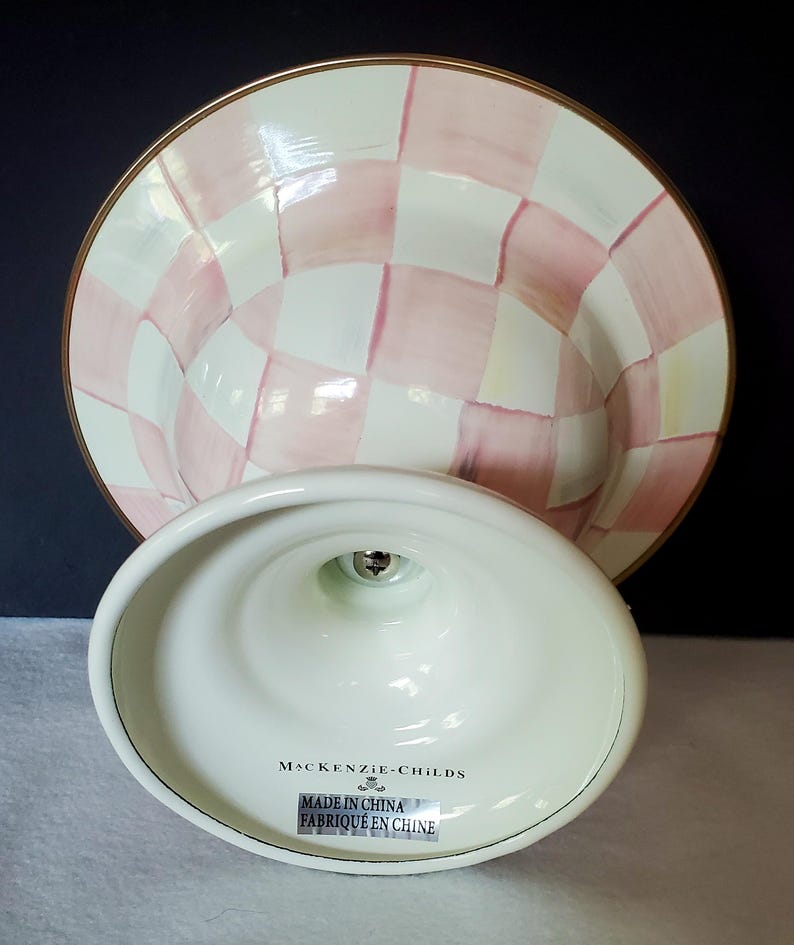 May include: A white and pink checkered cake stand with a gold rim. The base is white with the text "MacKenzie-Childs" and "Made in China / Fabriqu&eacute; en Chine". The stand is designed to hold a cake or other desserts.
