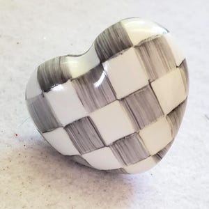 May include: Heart-shaped decorative object with a checkerboard pattern in shades of grey and white. The glossy surface reflects light, highlighting the hand-painted design. This unique piece could be used as a decorative accent or a functional item.