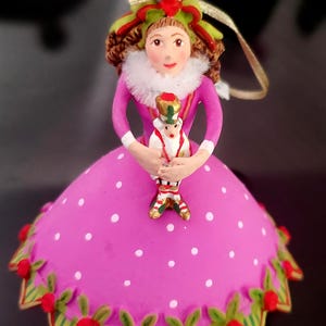 May include: A decorative Christmas ornament featuring a doll-like figure in a pink polka-dotted dress. The figure holds a smaller toy soldier. The ornament has a red heart on top and is trimmed with green and red accents.