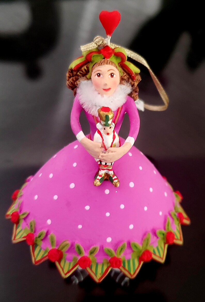 May include: A decorative Christmas ornament featuring a doll-like figure in a pink polka-dotted dress. The figure holds a smaller toy soldier. The ornament has a red heart on top and is trimmed with green and red accents.
