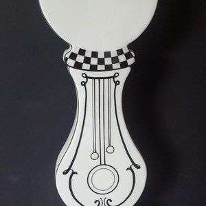May include: A tall, decorative white and black ceramic piece with a round top and a checkered pattern. The body features black lines and a decorative design. The base is square with a black border.