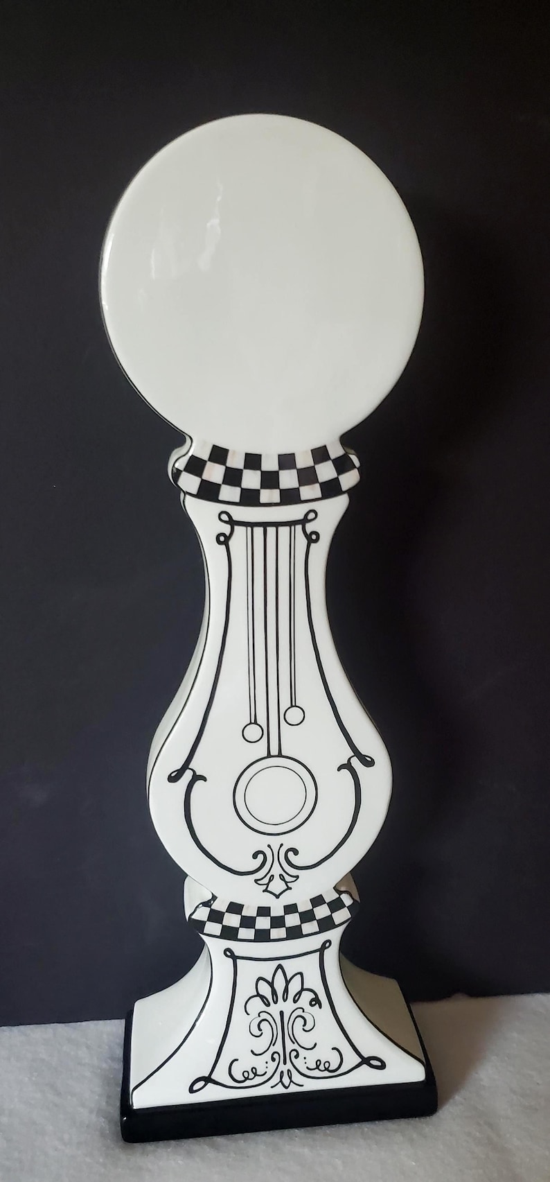 May include: A tall, decorative white and black ceramic piece with a round top and a checkered pattern. The body features black lines and a decorative design. The base is square with a black border.