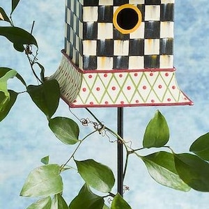 May include: A decorative birdhouse mounted on a thin black metal pole. The roof is light blue with red polka dots, topped with a colorful finial. The body features a black and white checkerboard pattern, while the base has a green diamond lattice.