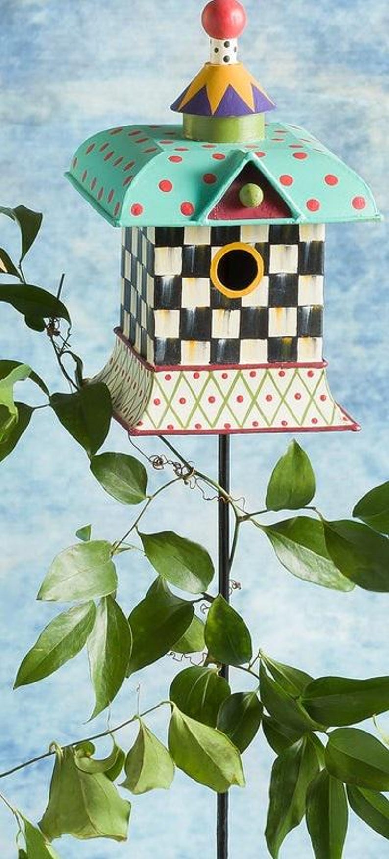 May include: A decorative birdhouse mounted on a thin black metal pole. The roof is light blue with red polka dots, topped with a colorful finial. The body features a black and white checkerboard pattern, while the base has a green diamond lattice.