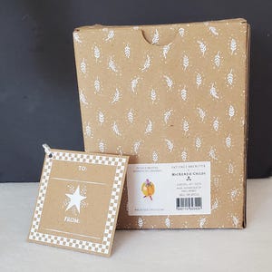 May include: A brown cardboard box and gift tag. The box has a white leaf pattern. The gift tag has a star design and the words "TO:" and "FROM:". The box has a label with the text "PATIENCE BREWSTER MACKENZIE-CHILDS".