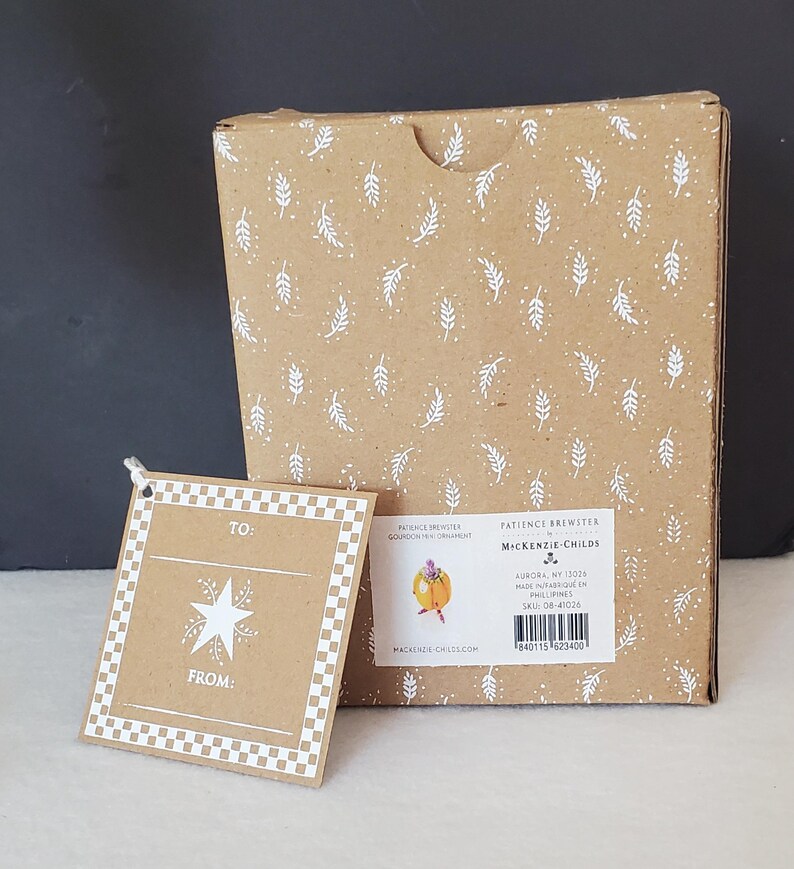May include: A brown cardboard box and gift tag. The box has a white leaf pattern. The gift tag has a star design and the words "TO:" and "FROM:". The box has a label with the text "PATIENCE BREWSTER MACKENZIE-CHILDS".