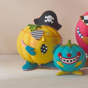 May include: A collection of pumpkin-shaped figurines. The central yellow pumpkin wears a pirate hat and eye patch. A teal pumpkin with a smiling face stands nearby. The figurines have colourful details and are set against a neutral background.