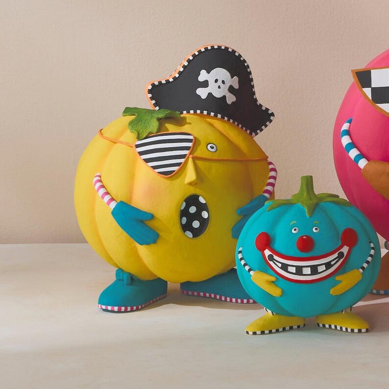 May include: A collection of pumpkin-shaped figurines. The central yellow pumpkin wears a pirate hat and eye patch. A teal pumpkin with a smiling face stands nearby. The figurines have colourful details and are set against a neutral background.