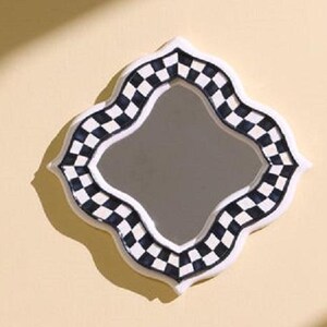 Puede incluir: Un espejo de pared con forma de cuatrifolio y un marco decorativo que presenta un patr&oacute;n de cuadros en blanco y negro. El espejo tiene una superficie central lisa y reflectante rodeada por un borde grueso con detalles blancos.