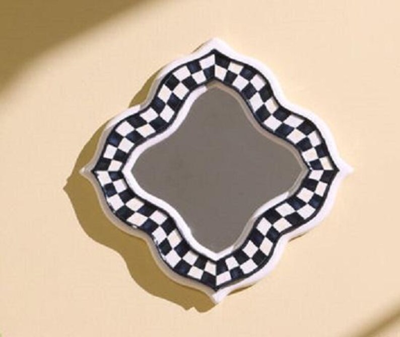 Puede incluir: Un espejo de pared con forma de cuatrifolio y un marco decorativo que presenta un patr&oacute;n de cuadros en blanco y negro. El espejo tiene una superficie central lisa y reflectante rodeada por un borde grueso con detalles blancos.