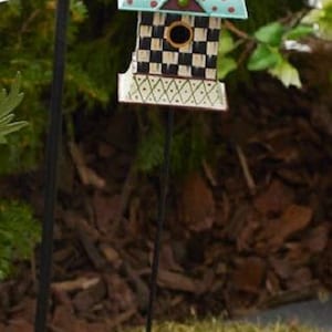 May include: A decorative metal birdhouse on a thin black stake featuring a black and white checkered pattern, a teal roof with red polka dots, and a small circular entrance. Above the birdhouse sits a pink scalloped metal bowl on a black stake.