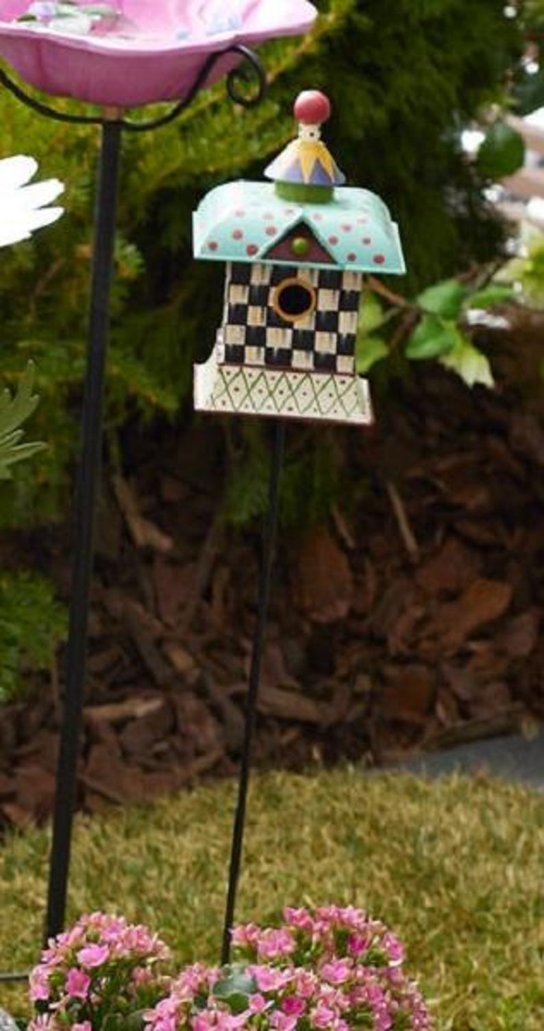 May include: A decorative metal birdhouse on a thin black stake featuring a black and white checkered pattern, a teal roof with red polka dots, and a small circular entrance. Above the birdhouse sits a pink scalloped metal bowl on a black stake.