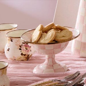 May include: A pink and white checkered pedestal serving bowl filled with cookies. The bowl has a gold rim and is surrounded by matching floral-patterned mugs and a vase. The table setting is on a pink tablecloth.