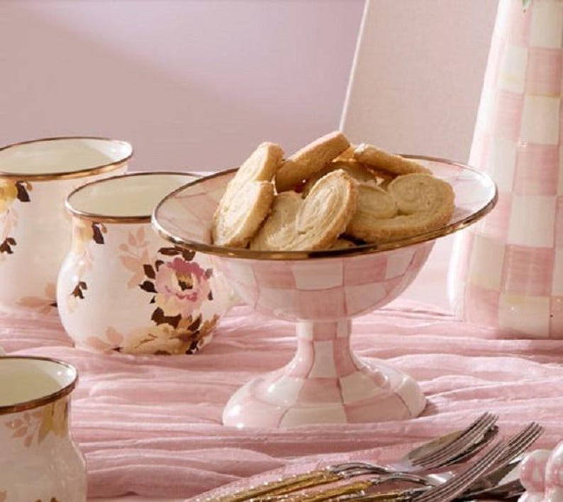 May include: A pink and white checkered pedestal serving bowl filled with cookies. The bowl has a gold rim and is surrounded by matching floral-patterned mugs and a vase. The table setting is on a pink tablecloth.