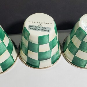 K&ouml;nnte beinhalten: Drei umgedrehte Metallbecher mit einem handgemalten gr&uuml;n-cremefarbenen Karomuster. Auf jedem Boden befindet sich der Schriftzug MacKenzie-Childs und Made in Viet Nam. Die Becher haben eine glatte, gl&auml;nzende Oberfl&auml;che und einen runden Rand.