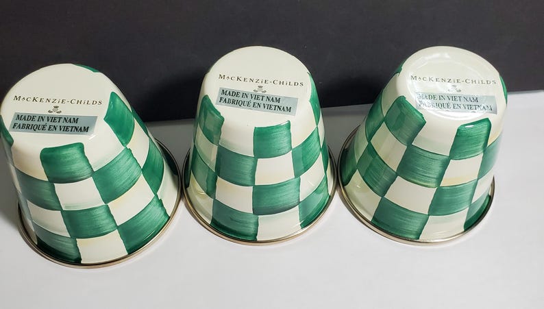 K&ouml;nnte beinhalten: Drei umgedrehte Metallbecher mit einem handgemalten gr&uuml;n-cremefarbenen Karomuster. Auf jedem Boden befindet sich der Schriftzug MacKenzie-Childs und Made in Viet Nam. Die Becher haben eine glatte, gl&auml;nzende Oberfl&auml;che und einen runden Rand.