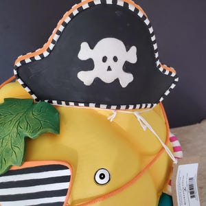 May include: A yellow decorative pumpkin wearing a black pirate hat with a white skull and crossbones. The hat has an orange and white striped trim. The pumpkin has a green leaf and orange detailing.