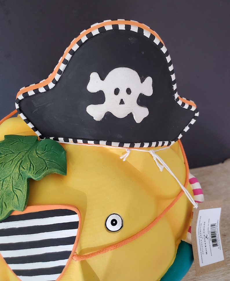 May include: A yellow decorative pumpkin wearing a black pirate hat with a white skull and crossbones. The hat has an orange and white striped trim. The pumpkin has a green leaf and orange detailing.
