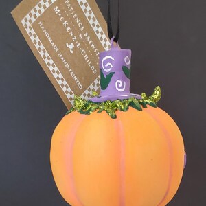 May include: A handmade, hand-painted pumpkin ornament with a witch hat and polka-dot shoes. The orange pumpkin has purple stripes and is topped with a purple hat with white swirls. The tag reads "PATIENCE BREWSTER" and "MacKenzie-Childs".