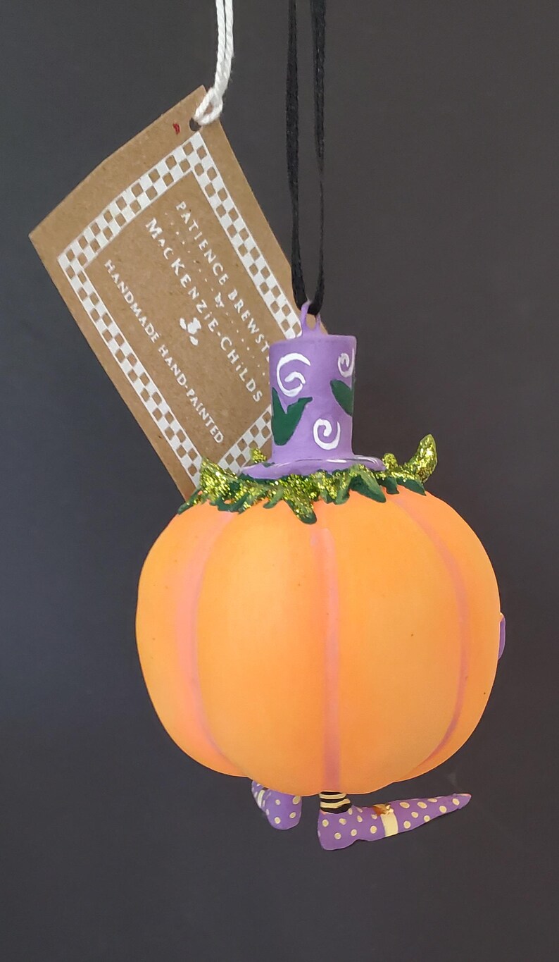 May include: A handmade, hand-painted pumpkin ornament with a witch hat and polka-dot shoes. The orange pumpkin has purple stripes and is topped with a purple hat with white swirls. The tag reads "PATIENCE BREWSTER" and "MacKenzie-Childs".