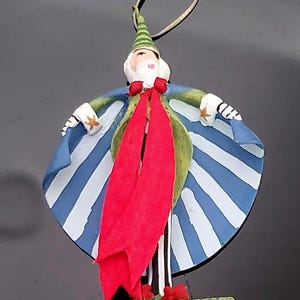 May include: A whimsical Christmas ornament depicting a figure with a white beard, a green and blue striped cape, and a red scarf. The ornament has a green hat and is standing on a green base. The ornament is made of painted wood.