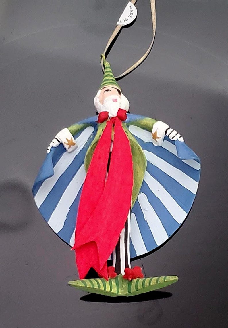 May include: A whimsical Christmas ornament depicting a figure with a white beard, a green and blue striped cape, and a red scarf. The ornament has a green hat and is standing on a green base. The ornament is made of painted wood.
