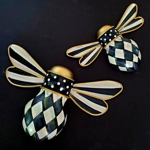 Puede incluir: Dos decoraciones de pared con forma de abeja que presentan cuerpos a cuadros blancos y negros, alas a rayas blancas y negras y cabezas doradas. La secci&oacute;n media de cada abeja es negra con lunares blancos y los bordes tienen acabado dorado.