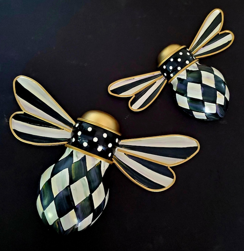 Puede incluir: Dos decoraciones de pared con forma de abeja que presentan cuerpos a cuadros blancos y negros, alas a rayas blancas y negras y cabezas doradas. La secci&oacute;n media de cada abeja es negra con lunares blancos y los bordes tienen acabado dorado.
