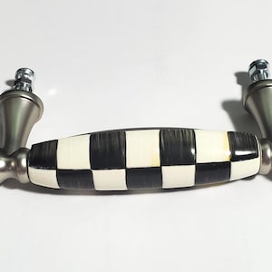 Może przedstawiać: Uchwyt meblowy z centralnym ceramicznym elementem w czarno-białą szachownicę, otoczony dwoma metalowymi wspornikami o wykończeniu ze szczotkowanego niklu z ozdobnymi, zaokrąglonymi i stopniowanymi zakończeniami, w zestawie dwie śruby montażowe.