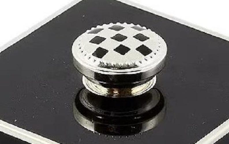 May include: A circular metal knob featuring a silver-toned finish with a repeating black diamond pattern on the top surface. The edge of the knob has a textured, beaded border, and the base is a smooth, reflective silver-toned metal cylinder.