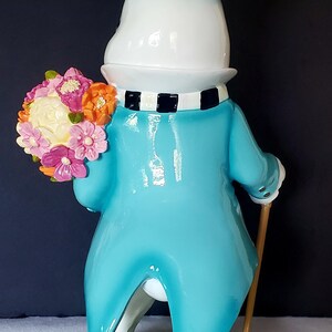 Puede incluir: Una figura caprichosa de un conejo blanco con un abrigo azul, sosteniendo un ramo de flores y un bast&oacute;n. El conejo lleva un sombrero de copa a cuadros blanco y negro y una bufanda a juego. La base es verde con flores.