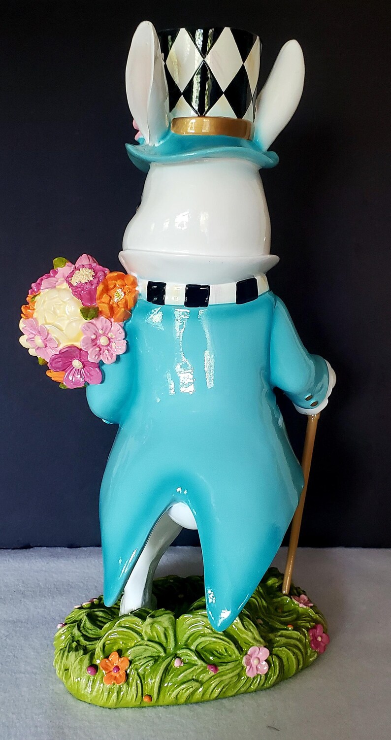 Puede incluir: Una figura caprichosa de un conejo blanco con un abrigo azul, sosteniendo un ramo de flores y un bast&oacute;n. El conejo lleva un sombrero de copa a cuadros blanco y negro y una bufanda a juego. La base es verde con flores.