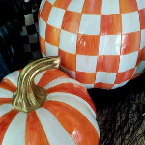 May include: Two decorative ceramic pumpkins. One pumpkin has a checkered pattern in orange and white. The other pumpkin has orange and white stripes. Both pumpkins have gold-colored stems. The pumpkins are set against a dark background.