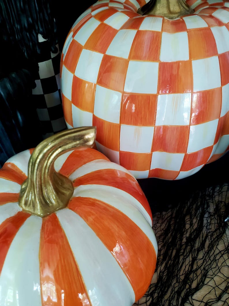 May include: Two decorative ceramic pumpkins. One pumpkin has a checkered pattern in orange and white. The other pumpkin has orange and white stripes. Both pumpkins have gold-colored stems. The pumpkins are set against a dark background.