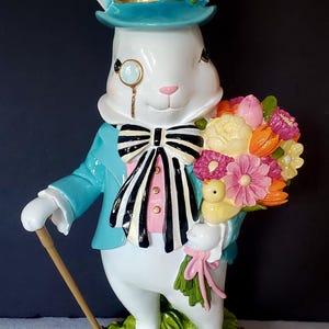 Puede incluir: Una caprichosa figura de conejo blanco, vestida con una chaqueta turquesa, chaleco rosa y pajarita en blanco y negro. Lleva un mon&oacute;culo y un sombrero de copa a cuadros, sosteniendo un ramo de flores coloridas y un bast&oacute;n. La figura est&aacute; sobre una base verde.