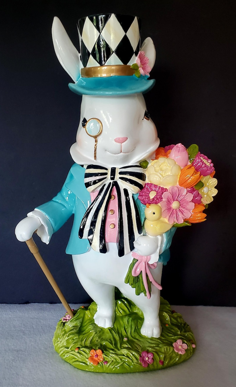 Puede incluir: Una caprichosa figura de conejo blanco, vestida con una chaqueta turquesa, chaleco rosa y pajarita en blanco y negro. Lleva un mon&oacute;culo y un sombrero de copa a cuadros, sosteniendo un ramo de flores coloridas y un bast&oacute;n. La figura est&aacute; sobre una base verde.