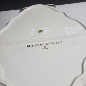 May include: The underside of a white ceramic serving tray with a scalloped edge. The text MACKENZIE-CHILDS is printed in black lettering above a small gold emblem. A circular yellow sticker with the text MADE IN CHINA is visible near the top.
